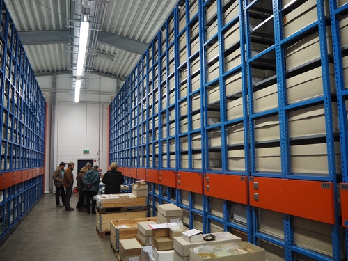 Photo showing the interior of a large storage facility with a high ceiling.
The space is dominated by massive, floor-to-ceiling blue metal industrial shelving units that line both sides of a central aisle. The shelves are densely packed. A small group of people are gathered around a makeshift workstation in the aisle.