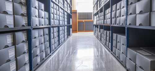 An eye-level, wide shot shows a narrow aisle between tall, blue metal shelving units in an archive or storage facility. The shelves are packed with hundreds of uniform, grey archival boxes, each featuring a white label on the side. The floor is a polished, reflective light grey, mirroring the bright overhead lighting. In the background, a wooden doorway and a dark grey partition are visible at the end of the long corridor of shelves.