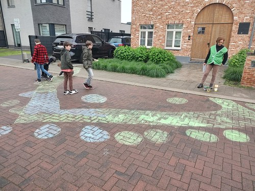 A street-level colour photo shows a group of children filling in a large, L-shaped chalk outline on a brick-paved street with green and blue chalk. Several circular shapes are also filled in with the same colors around the main shape.
A woman in a green safety vest stands to the right, watching the children. Boxes of chalk are on the ground near her feet. In the background, there's a brick building with a large wooden door, two parked cars, and a modern house.